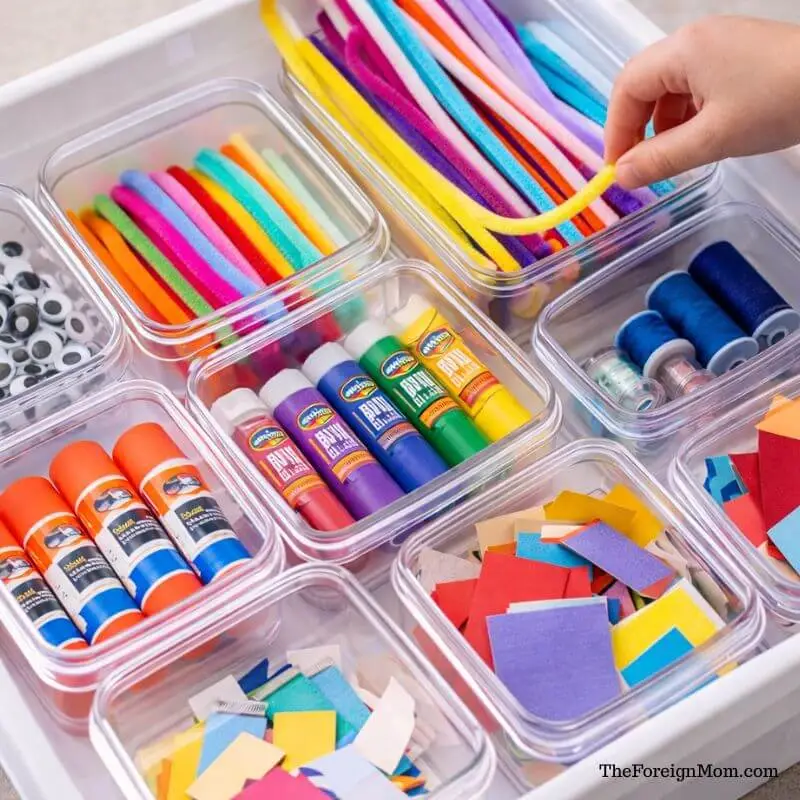 Clear containers organizing kids’ art supplies including googly eyes, pipe cleaners, glue sticks, paint tubes, and paper scraps for easy access.
