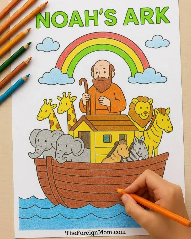 A child's hand coloring a Noah's Ark Catholic coloring page with animals, rainbow, and Noah on a wooden table.