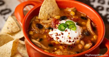 Bowl of hearty chicken taco soup topped with sour cream, jalapenos, and tortilla chips. Served in a cozy crockpot-style presentation.