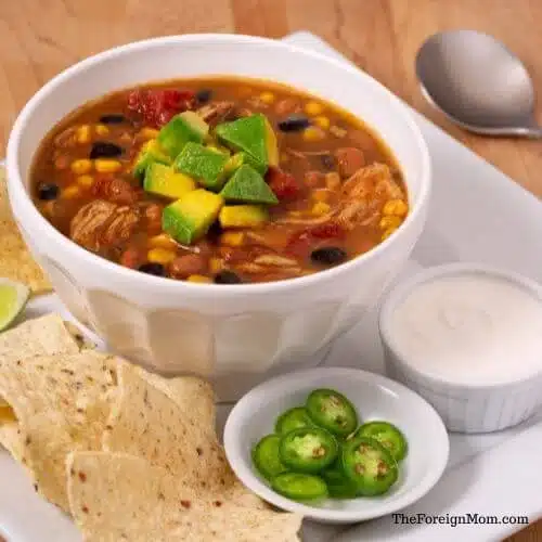 Bowl of chicken taco soup topped with avocado and served with tortilla chips and sliced jalapenos.