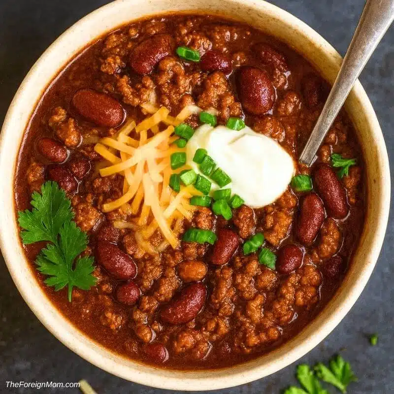 A hearty bowl of classic beef chili topped with sour cream, sliced green onions, and melted cheddar cheese, served in a ceramic bowl with fresh parsley.