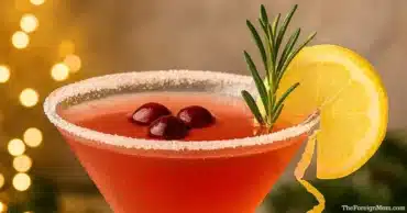 A festive cranberry lemon drop martini served in a chilled martini glass, garnished with fresh cranberries, a lemon wheel, and a rosemary sprig against a warm holiday background.