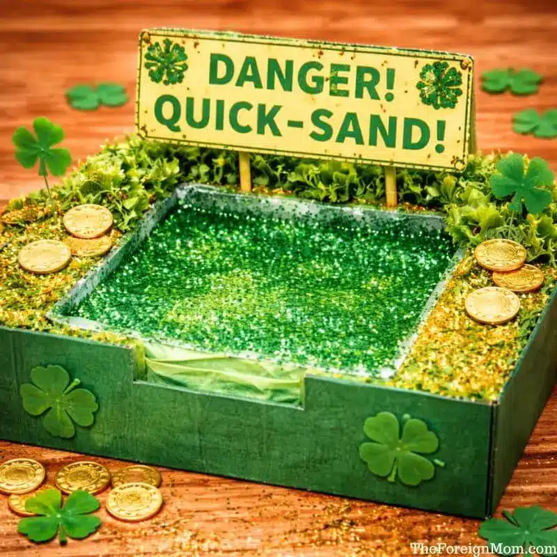 Glitter glue quick-sand leprechaun trap made from a cardboard box with green glitter glue, gold coins, and shamrock decorations for St. Patrick’s Day.