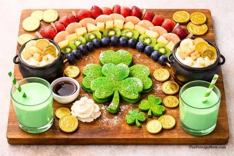 St. Patrick’s Day breakfast board with shamrock pancakes, fruit rainbow, yogurt cups, green milk, and gold chocolate coins on a wooden tray.