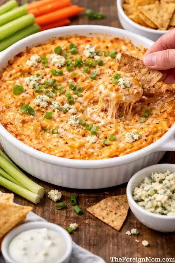 Creamy healthy buffalo chicken dip being scooped with a tortilla chip, surrounded by celery and carrot sticks, perfect for game day.