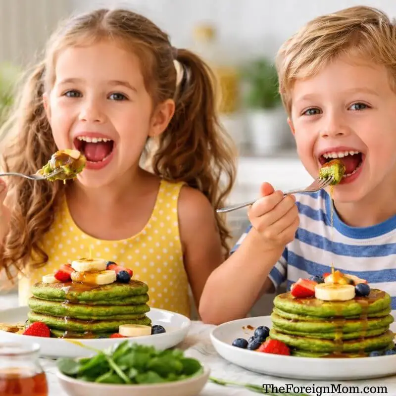 Two kids eating healthy green pancakes made with spinach and banana, topped with fruit and maple syrup at the breakfast table.