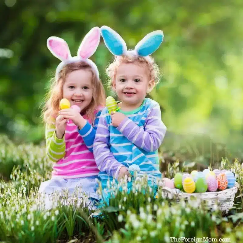 Two preschool kids wearing bunny ears sitting in grass holding colorful Easter eggs during an outdoor egg hunt.