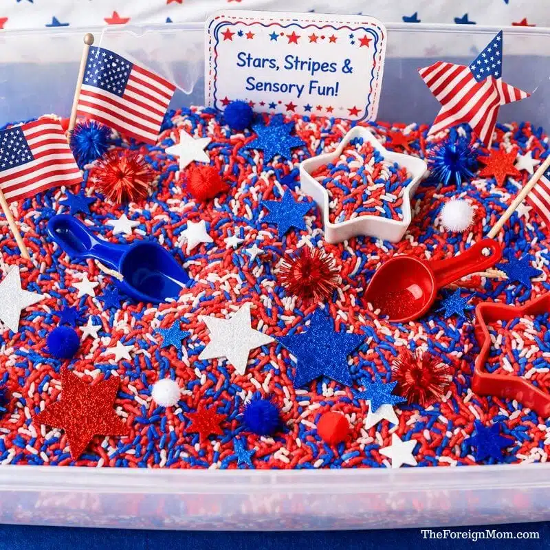 Red white and blue patriotic sensory bin with colored rice, stars, scoops, and mini American flags for kids sensory play.