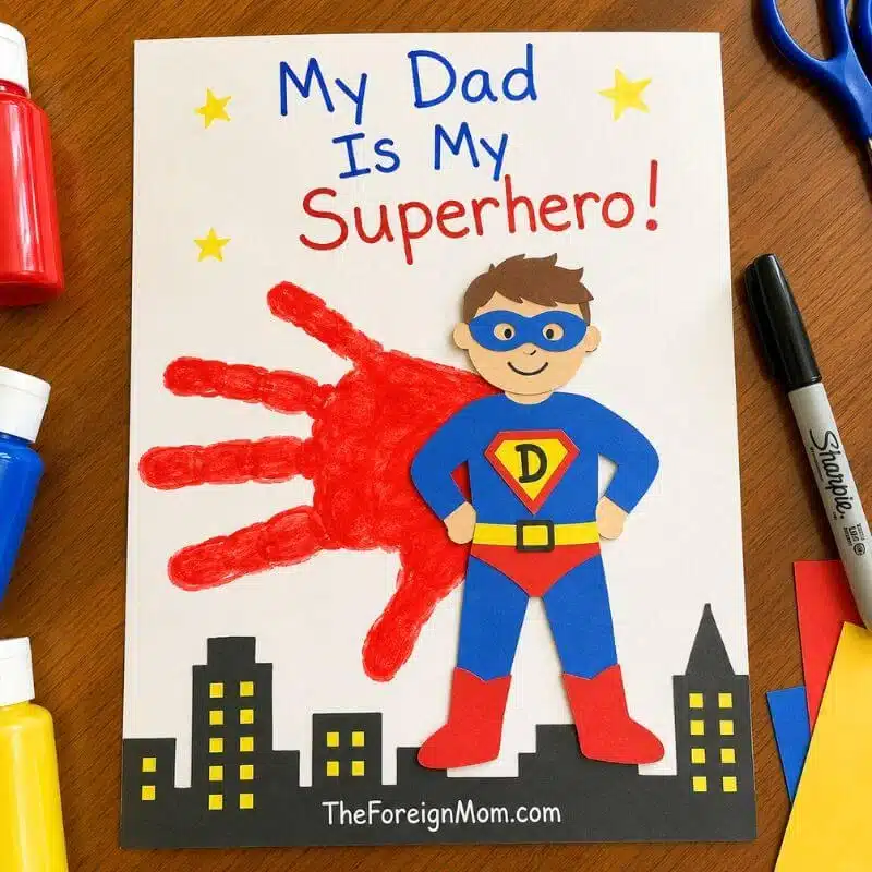 Father’s Day superhero handprint craft with red paint, paper superhero, and the words My Dad Is My Superhero.