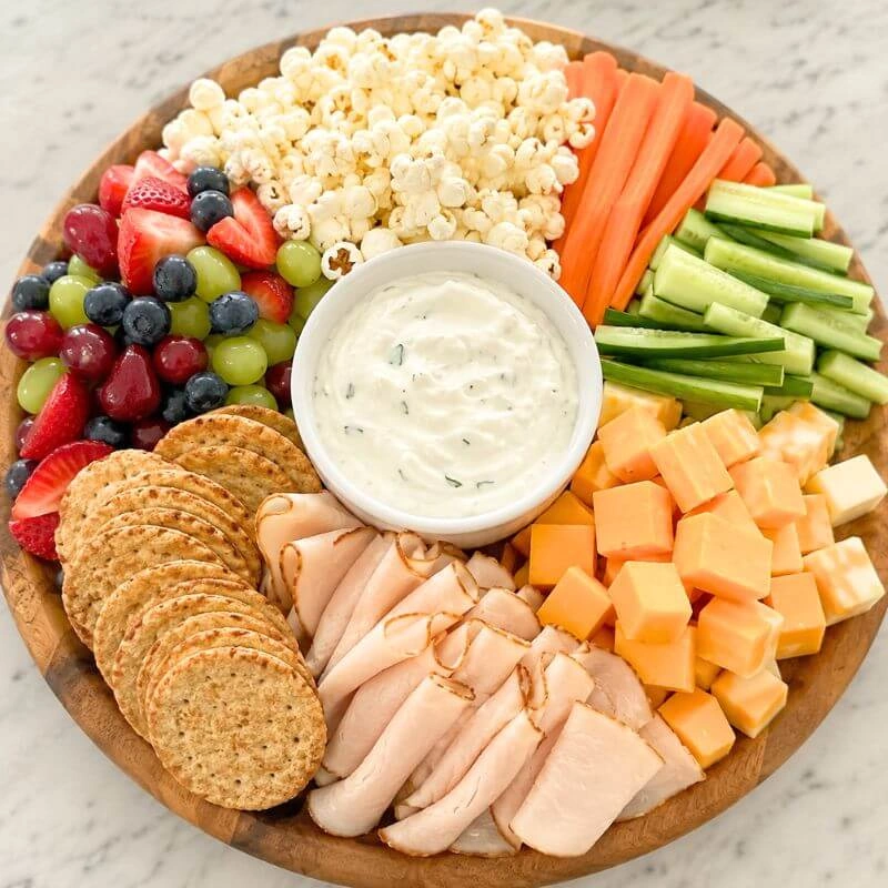 DIY snack board with fresh fruit, sliced vegetables, popcorn, cheese cubes, yogurt dip, whole grain crackers, and turkey slices.