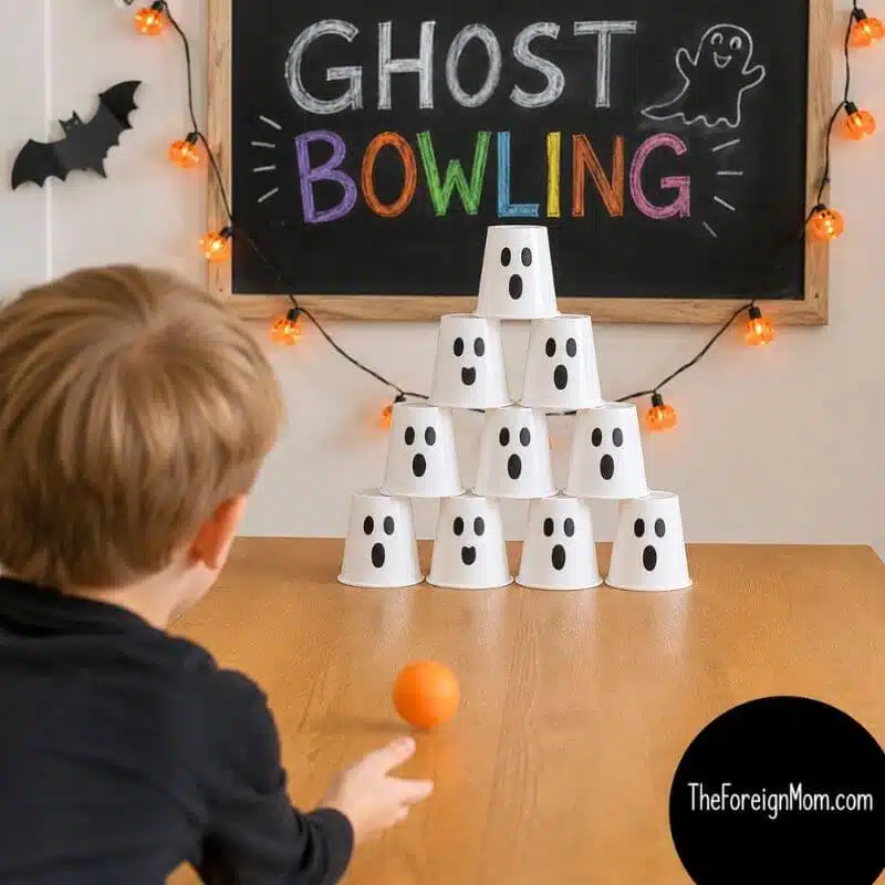 Kid playing a ghost bowling Halloween game with white plastic cups decorated with ghost faces and a small orange ball.