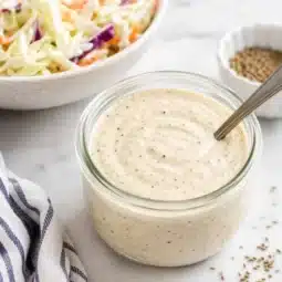 Homemade Marie’s coleslaw dressing copycat in a glass jar with creamy coleslaw in the background.