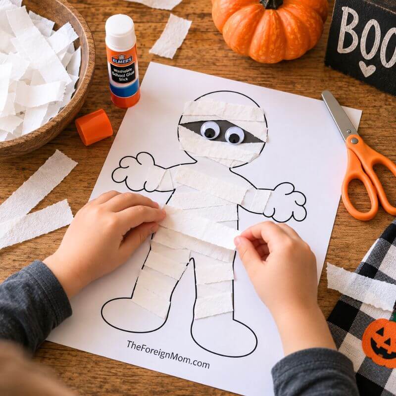 Child gluing white paper strips onto a mummy template for an easy Halloween craft.