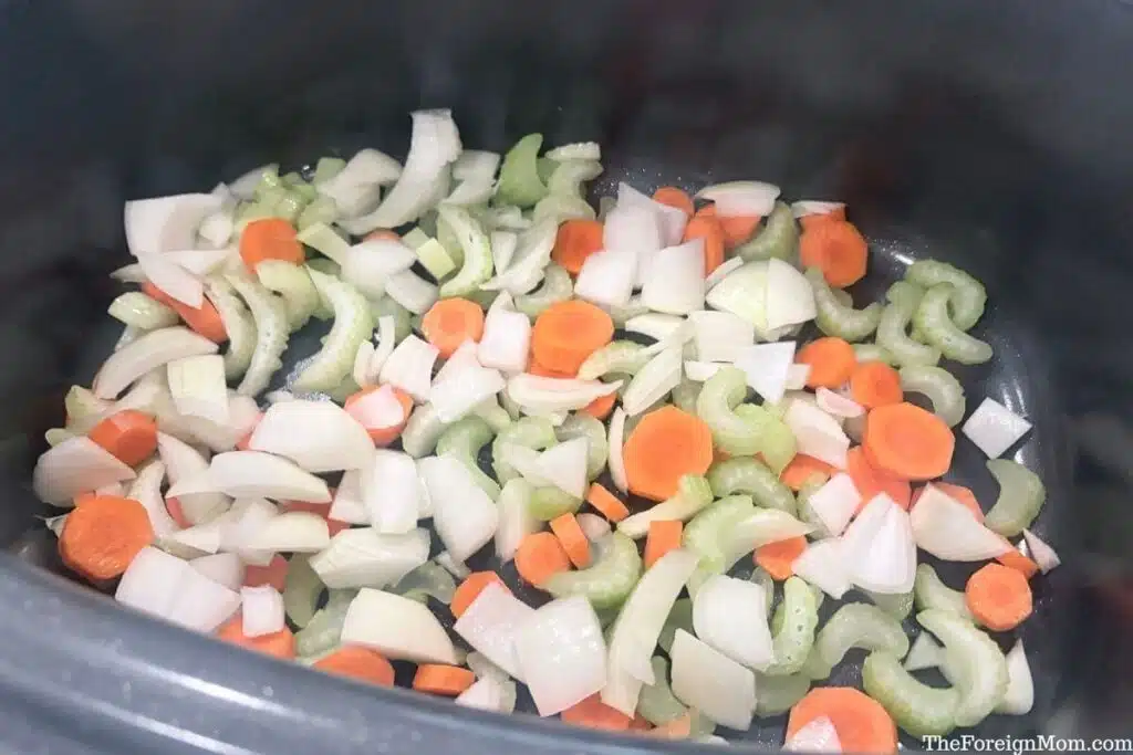 Chopped carrots, onion, and celery spread across the bottom of a slow cooker.