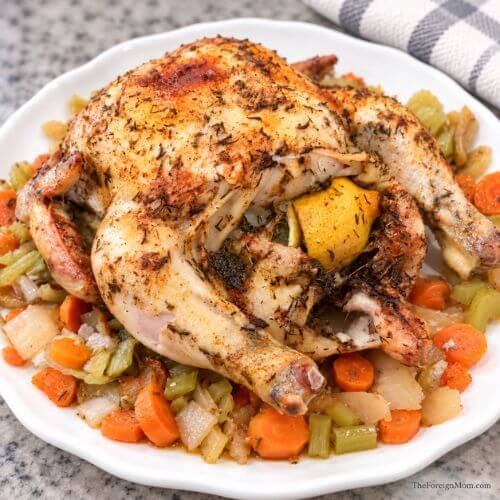 Lemon herb whole chicken with a lemon in the cavity, served with carrots, celery, and onions on a white platter.