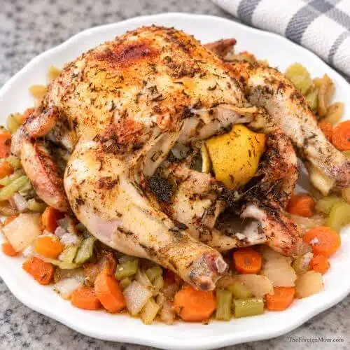 Lemon herb whole chicken with a lemon in the cavity, served with carrots, celery, and onions on a white platter.