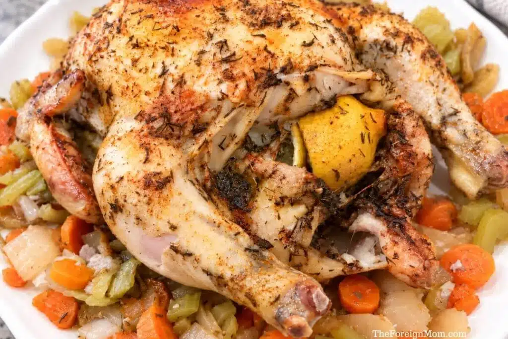 Lemon herb whole chicken with browned skin on a white platter surrounded by carrots, celery, and onions.