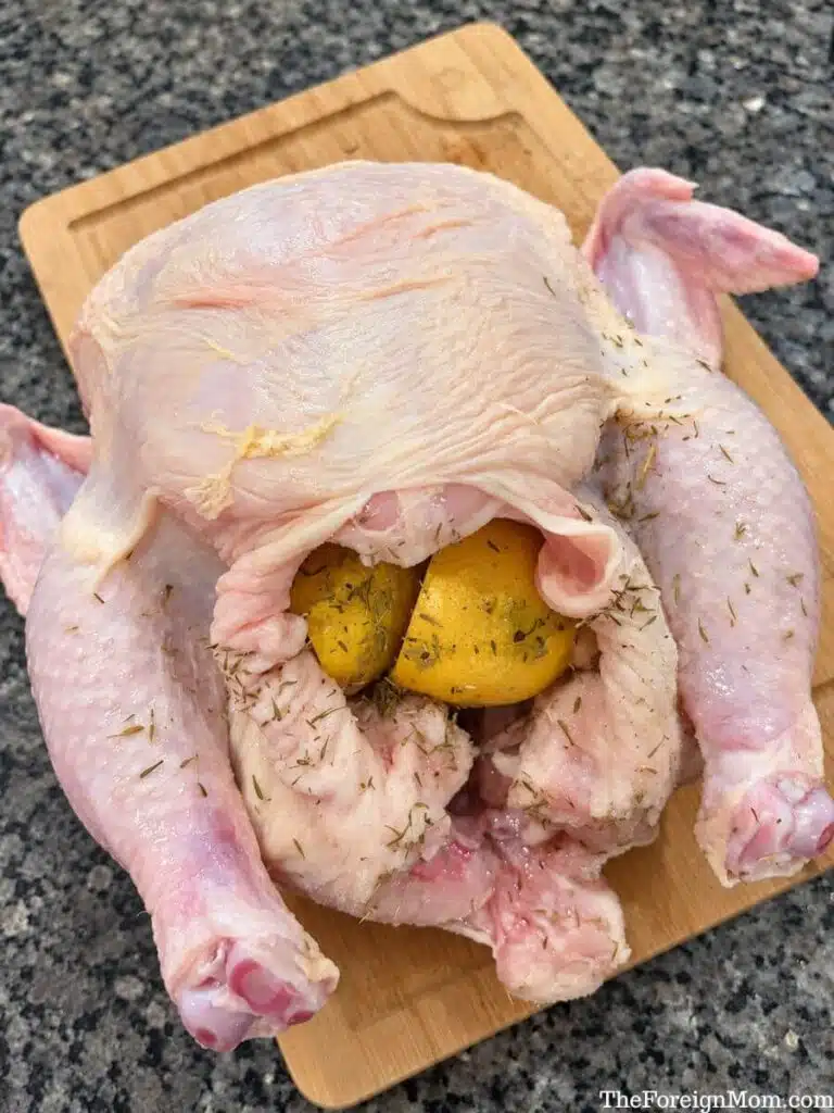 Raw whole chicken with lemon halves and thyme inside the cavity on a wooden cutting board.