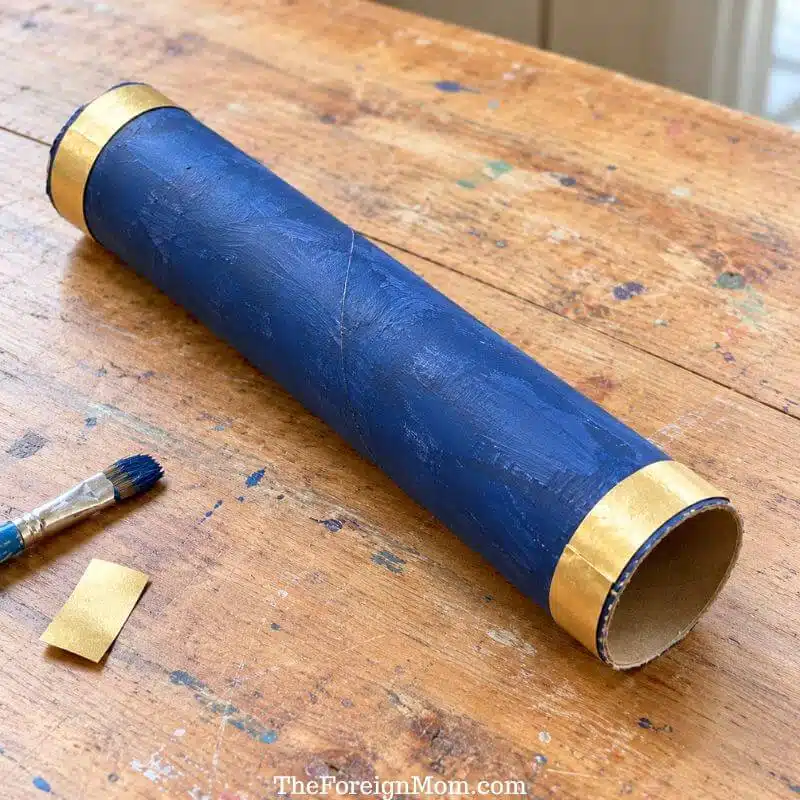 Navy blue cardboard tube telescope with gold paper bands on a wooden craft table.