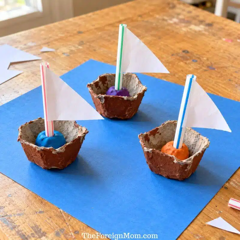 Three brown egg carton boats with straw masts and white paper sails on blue construction paper.