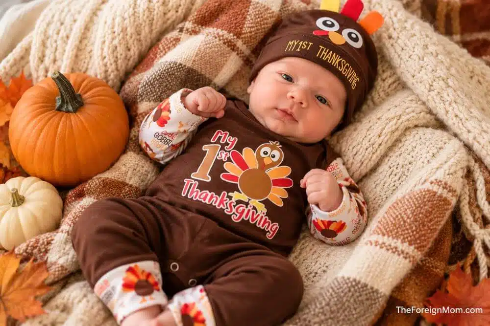 Newborn baby boy wearing a My First Thanksgiving turkey outfit beside pumpkins and cozy fall blankets.