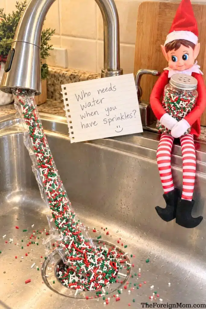 Elf on the Shelf holding Christmas sprinkles beside a kitchen faucet with a sprinkle-filled plastic sleeve and a handwritten “Who needs water when you have sprinkles?” note.