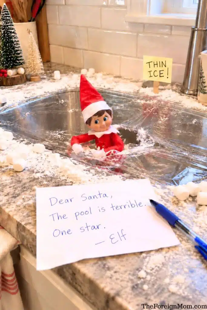 Funny Elf on the Shelf idea featuring an elf falling through plastic wrap over a kitchen sink decorated with fake snow, a thin ice warning sign, and a silly note to Santa.