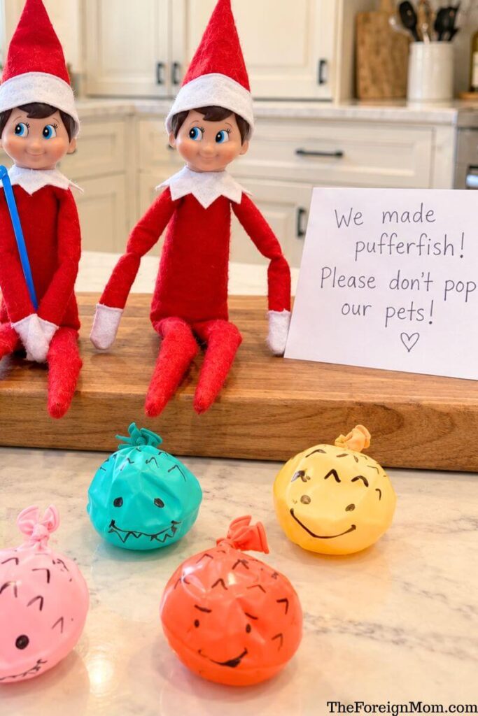 Funny Elf on the Shelf idea featuring two elves with colorful homemade balloon pufferfish pets decorated with silly faces and a handwritten note asking not to pop them.
