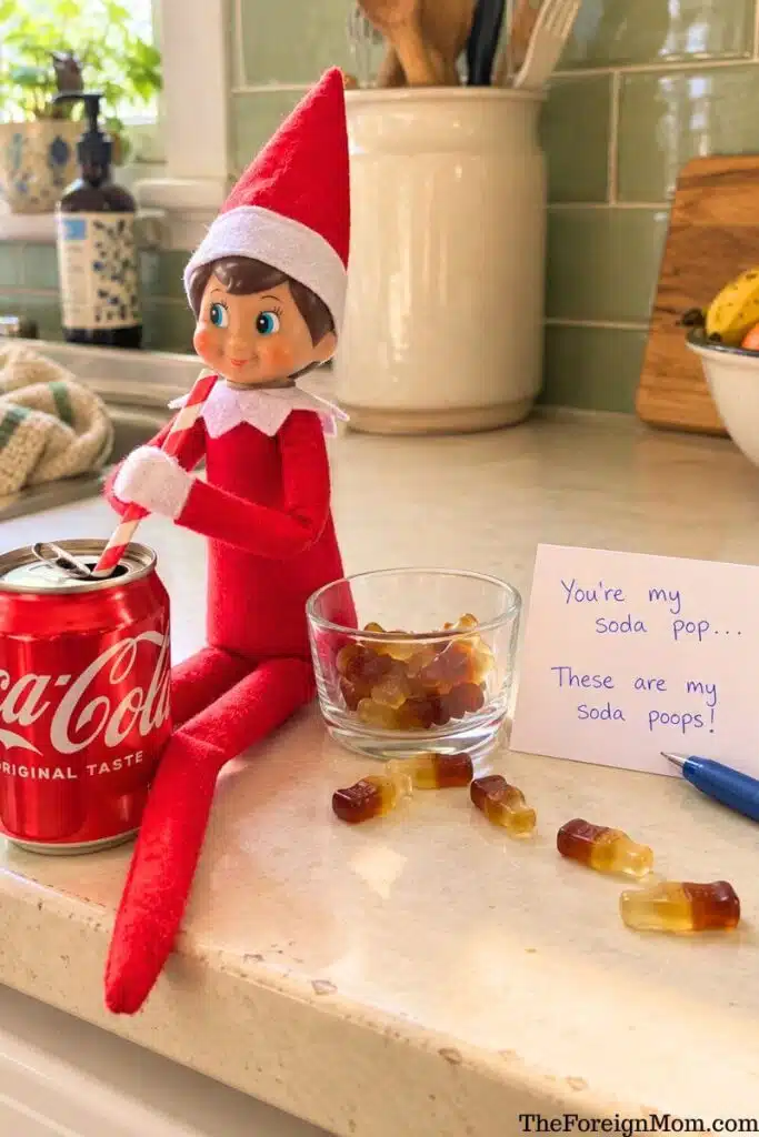 Funny Elf on the Shelf idea featuring an elf sitting beside a Coca-Cola can with gummy cola bottles and a silly soda poop note.
